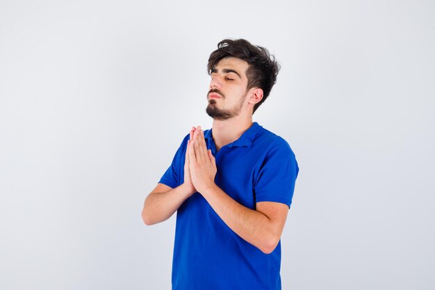 Giovane ragazzo in piedi in posa di preghiera in maglietta blu e sembra serio. vista frontale.