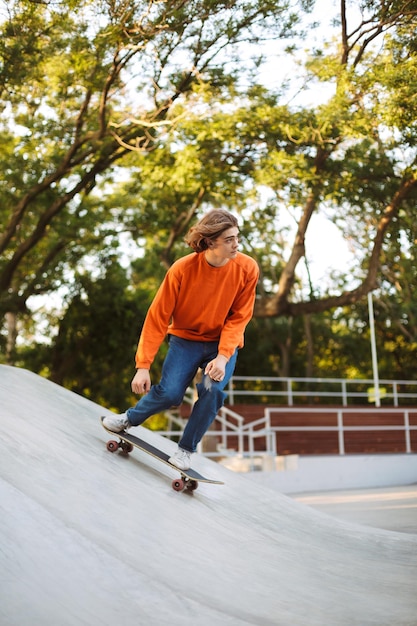 Giovane ragazzo in pullover arancione che fa skateboard e pratica acrobazie mentre trascorre del tempo nel moderno skatepark