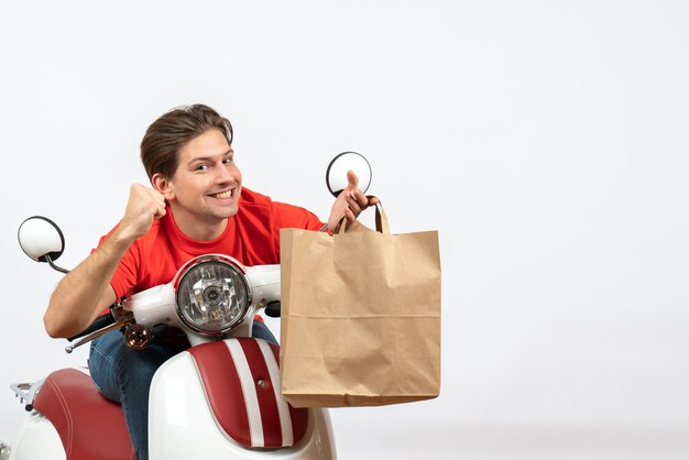 Giovane ragazzo orgoglioso sorridente del corriere in uniforme rossa che si siede sul motorino che tiene il sacchetto di carta sulla parete bianca