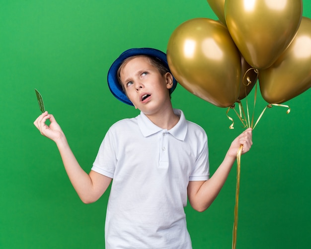 giovane ragazzo slavo all'oscuro con cappello da festa blu che tiene palloncini di elio e carta di credito guardando in alto isolato sulla parete verde con spazio di copia