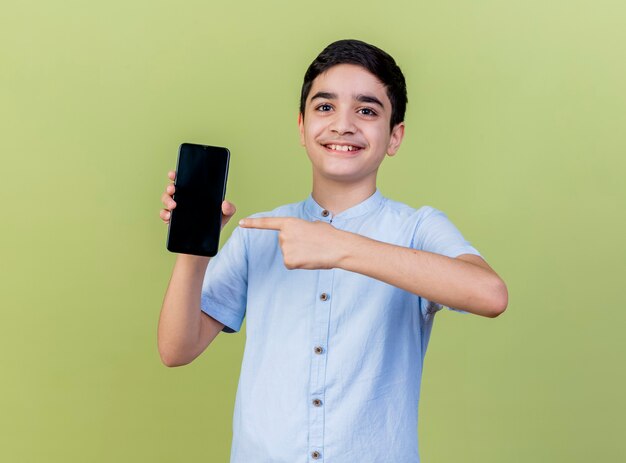 Giovane ragazzo sorridente che mostra e che indica al telefono cellulare che esamina la parte anteriore isolata sulla parete verde oliva