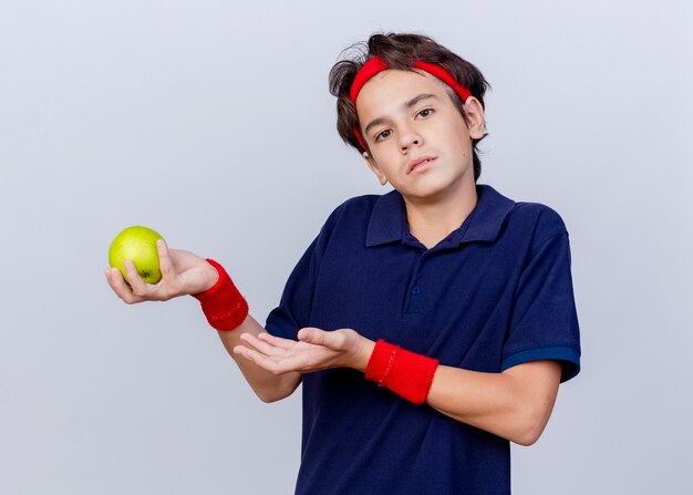 Giovane ragazzo sportivo bello all'oscuro che indossa la fascia e i braccialetti con le parentesi graffe dentali che tengono e che indica con la mano alla mela isolata sulla parete bianca