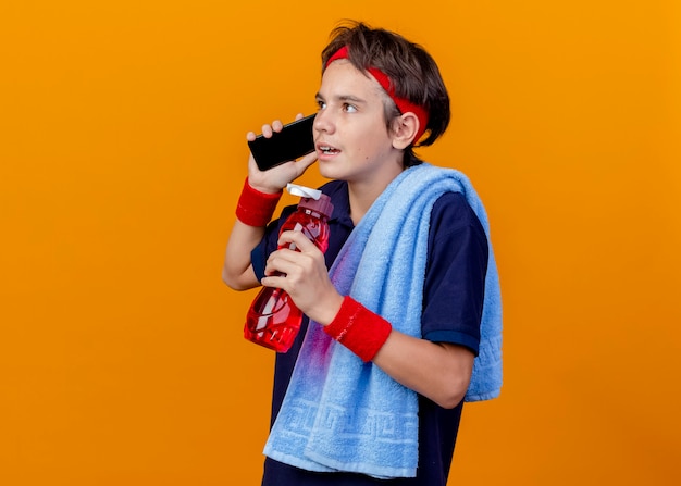 Giovane ragazzo sportivo bello che indossa fascia e braccialetti con bretelle dentali e asciugamano sulla spalla tenendo la bottiglia d'acqua parlando al telefono guardando dritto isolato sulla parete arancione con lo spazio della copia