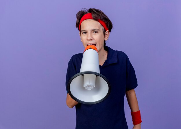 Giovane ragazzo sportivo bello che indossa la fascia e i braccialetti con le parentesi graffe dentali guardando la parte anteriore parlando da altoparlante isolato sulla parete viola