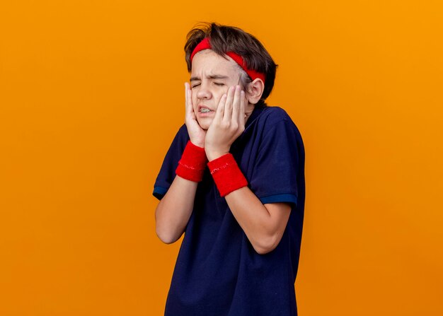 Giovane ragazzo sportivo bello dolorante che indossa la fascia e i braccialetti con le parentesi graffe dentali che tengono le mani sul viso con gli occhi chiusi isolati sulla parete arancione