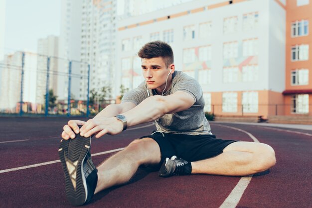 Giovane ragazzo sportivo in allenamento al mattino sullo stadio. Indossa una maglietta grigia, pantaloncini neri, fa stretching.