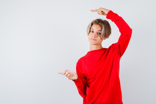 Giovane ragazzo teenager che indica a sinistra in maglione rosso e che sembra fiducioso, vista frontale.