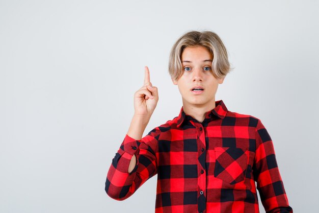 Giovane ragazzo teenager che indica in su in camicia controllata e che sembra perplesso. vista frontale.