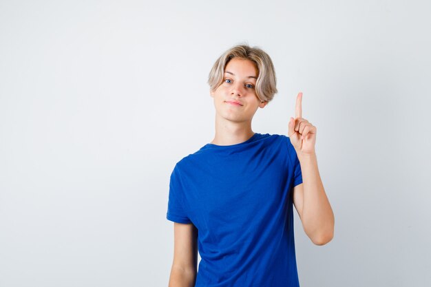 Giovane ragazzo teenager che indica in su in maglietta blu e che sembra fiducioso, vista frontale.
