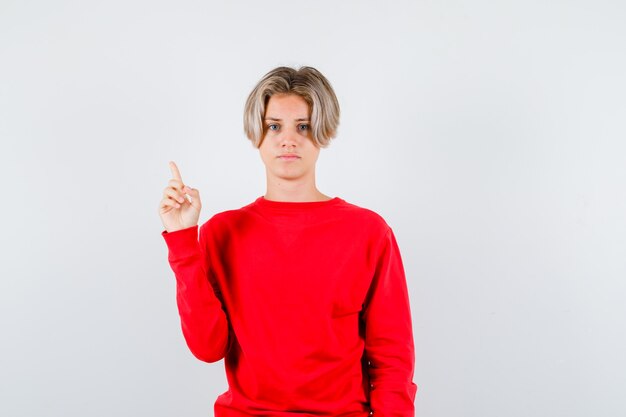 Giovane ragazzo teenager che indica in su in maglione rosso e che sembra astuto. vista frontale.