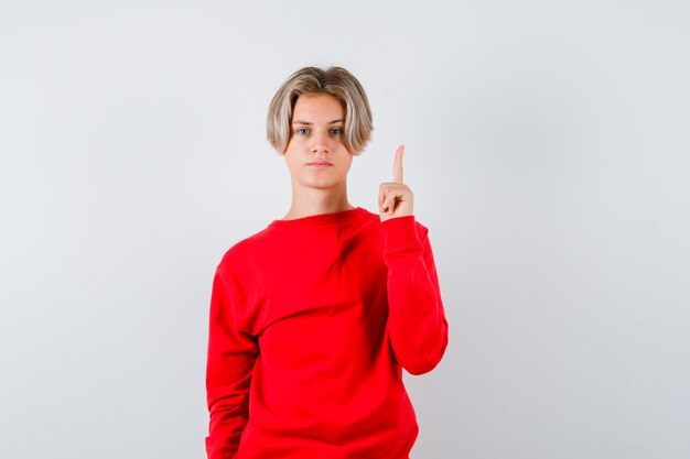 Giovane ragazzo teenager che indica in su in maglione rosso e che sembra serio. vista frontale.