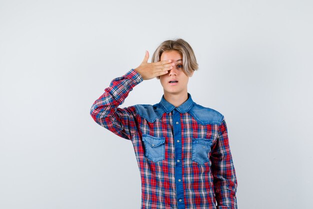 Giovane ragazzo teenager con la mano sull'occhio in camicia a quadri e guardando domandato. vista frontale.