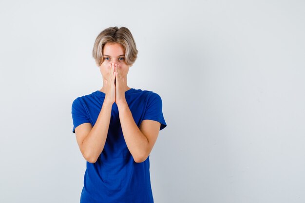 Giovane ragazzo teenager con le mani nel gesto di preghiera in maglietta blu e guardando speranzoso. vista frontale.