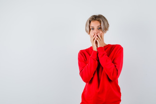 Giovane ragazzo teenager con le mani sulla bocca in maglione rosso e sembra ansioso. vista frontale.