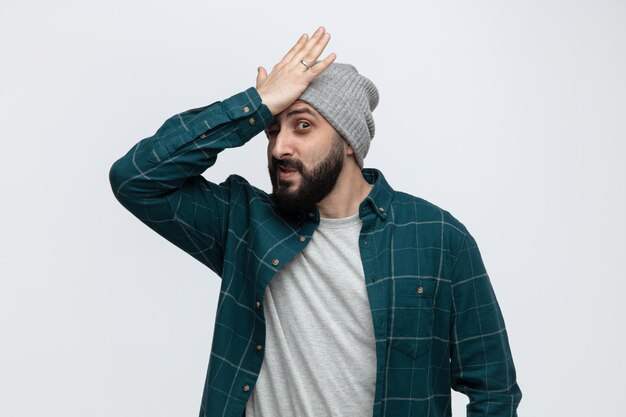 Giovane rammaricato che indossa il cappello invernale tenendo la mano sulla testa guardando la fotocamera isolata su sfondo bianco