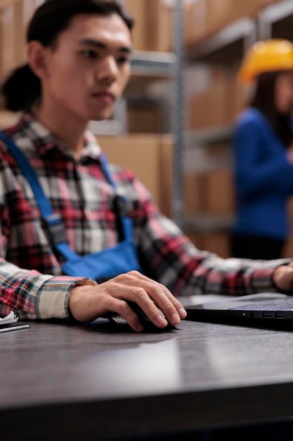 Giovane responsabile logistico asiatico che analizza la fornitura di merci sul laptop. Magazziniere che esegue la gestione dell'inventario sul computer mentre è seduto alla scrivania con il fuoco selettivo da vicino a portata di mano