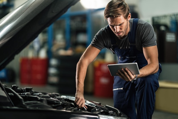 Giovane riparatore di auto che lavora sul motore dell'auto mentre utilizza la tavoletta digitale in officina