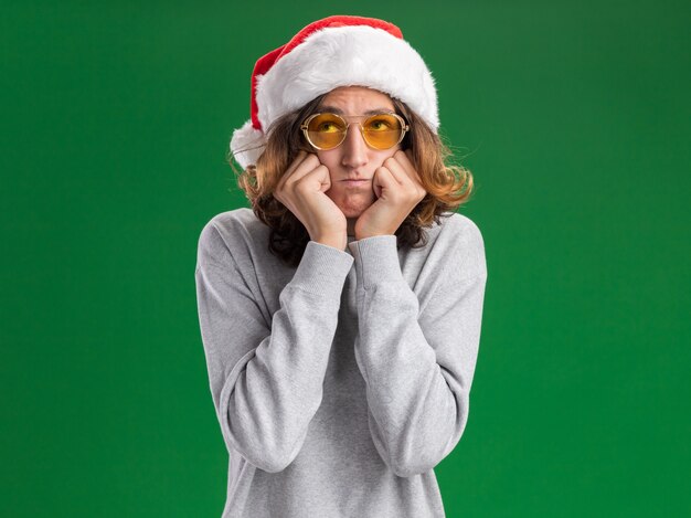 Giovane risentito che indossa il cappello di Babbo Natale e occhiali gialli che guardano le guance che soffiano in piedi sul muro verde