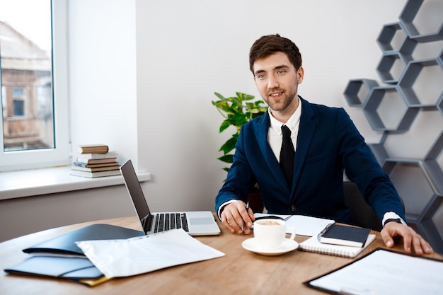 Giovane riuscito uomo d'affari che si siede nel luogo di lavoro, fondo dell'ufficio.