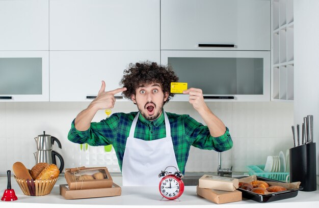 Giovane scioccato in piedi dietro il tavolo con vari pasticcini e mostrando la carta di credito nella cucina bianca