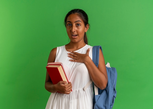 Giovane scolara sorpresa che porta il libro della tenuta della borsa posteriore con il taccuino e che mette la mano sul petto isolata sul verde