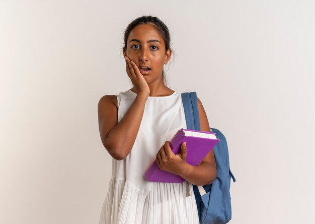 Giovane scolara sorpresa che porta il libro della tenuta della borsa posteriore e che mette la mano sulla guancia su bianco