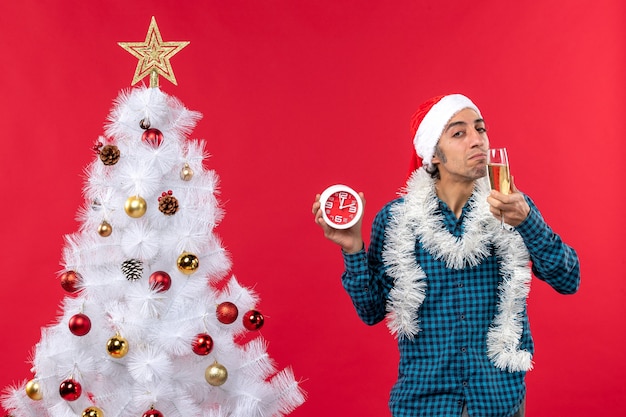 Giovane sicuro con cappello di Babbo Natale e degustazione di un bicchiere di vino e orologio in piedi vicino all'albero di Natale