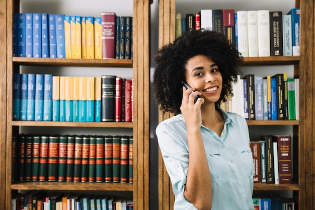 Giovane signora afroamericana sorridente che parla sullo smartphone vicino ai libri