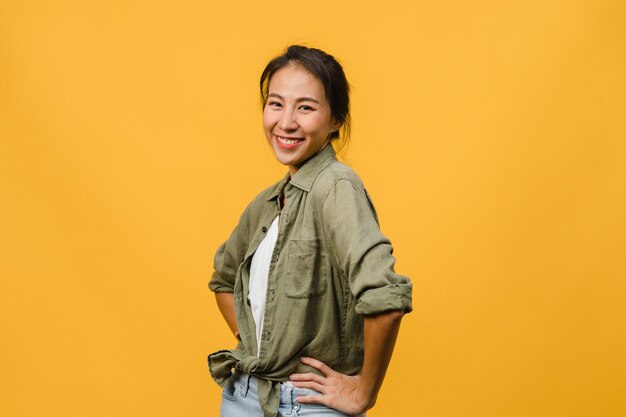 Giovane signora asiatica con un'espressione positiva, sorride ampiamente, vestita con abiti casual sul muro giallo. La donna felice adorabile felice si rallegra del successo. Concetto di espressione facciale.