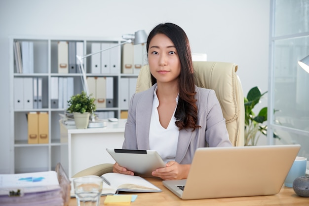 Giovane signora asiatica di affari che posa nell'ufficio con la compressa davanti al computer portatile
