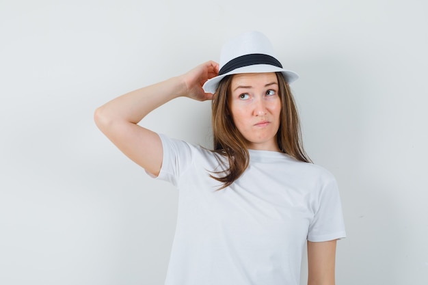 Giovane signora che graffia la testa in cappello bianco della maglietta e che sembra esitante