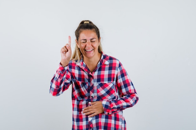 Giovane signora che indica in su in camicia controllata e che sembra felice. vista frontale.