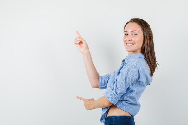 Giovane signora che indica le dita in su in camicia blu, pantaloni e sguardo allegro