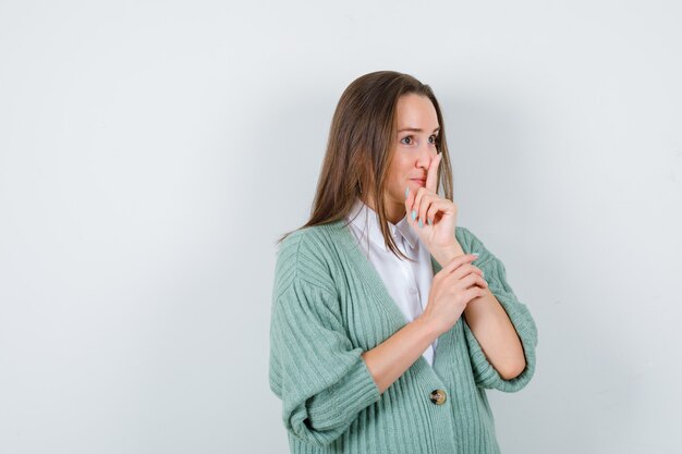 Giovane signora che mostra gesto di silenzio in camicia, cardigan e guardando fiducioso, vista frontale.