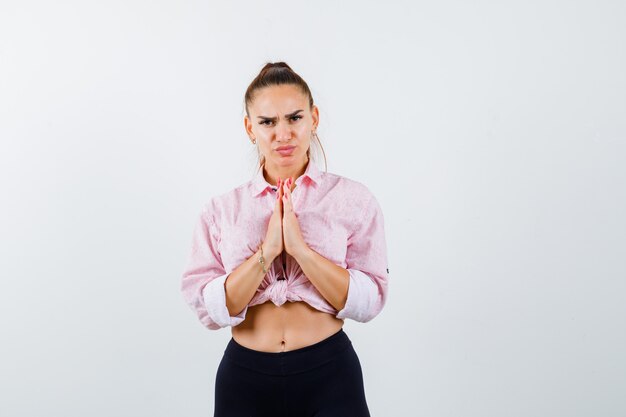 Giovane signora che mostra il gesto di namaste in camicia, pantaloni e sguardo pieno di speranza