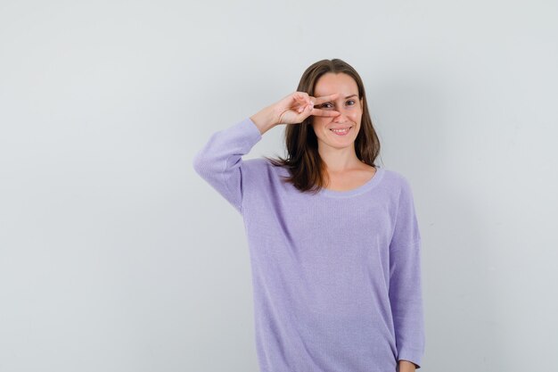 Giovane signora che mostra il segno di V sull'occhio in camicia casuale e che sembra felice. vista frontale.