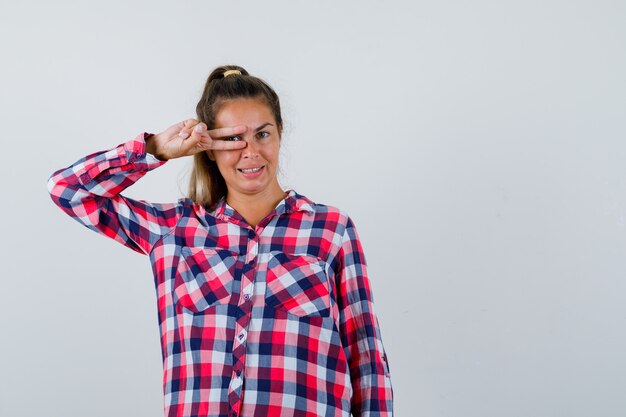 Giovane signora che mostra il segno di V sull'occhio in camicia controllata e che sembra felice, vista frontale.