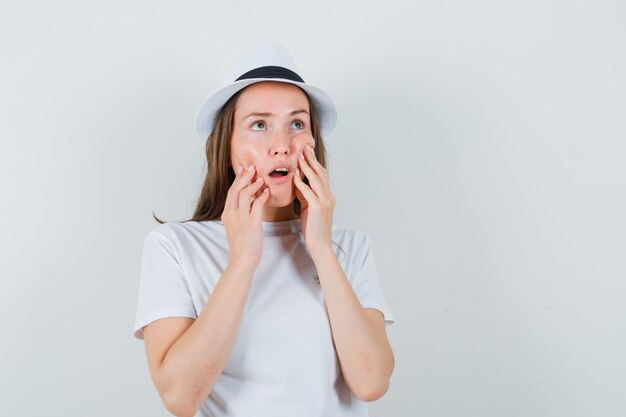 Giovane signora che preme le dita sulle guance in cappello bianco della maglietta e che sembra confusa