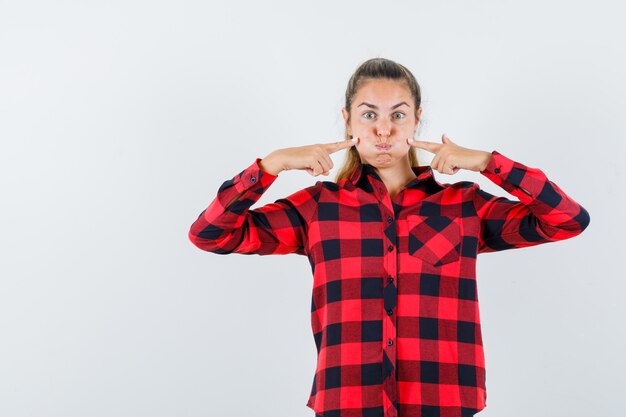 Giovane signora che preme le dita sulle guance soffiate in camicia a quadri e sembra divertente