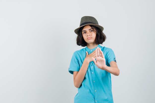 Giovane signora che rifiuta qualcosa in camicia blu, cappello e sguardo grato.