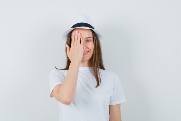 Giovane signora che tiene la mano sull'occhio in cappello bianco della maglietta e che sembra allegro