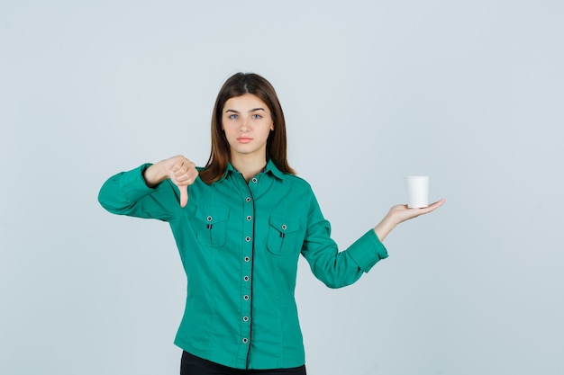 Giovane signora che tiene la tazza di caffè di plastica mentre mostra il pollice giù in camicia e sembra insoddisfatta, vista frontale.