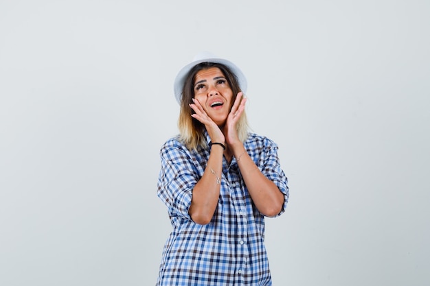 Giovane signora che tiene le mani sulle guance in cappello della camicia controllato e che sembra carina