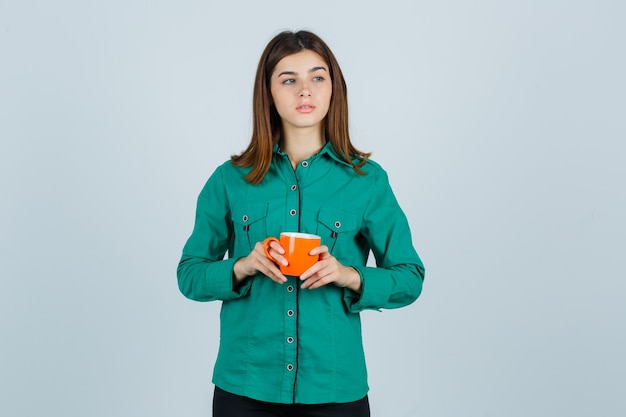 Giovane signora che tiene tazza di tè arancione in camicia e guardando concentrato, vista frontale.