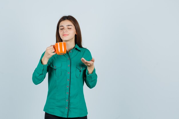 Giovane signora che tiene una tazza di tè arancione, mostrando qualcosa in camicia e guardando pacifica. vista frontale.