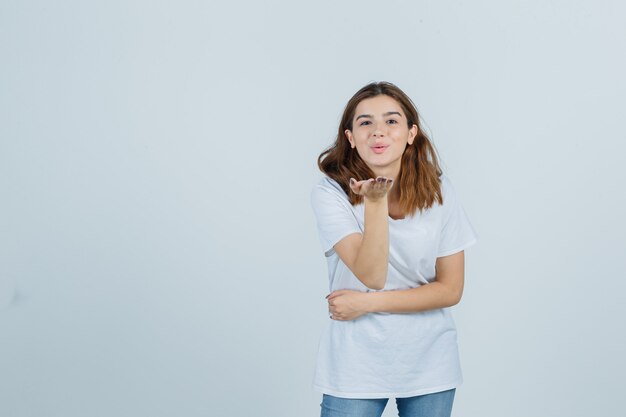 Giovane signora che trasmette bacio con la mano in maglietta, jeans e che sembra carina, vista frontale.