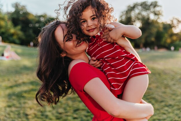 Giovane signora dai capelli castani in rosso che tiene sua figlia. Elegante modello femminile di trascorrere del tempo nel parco con il suo bambino.