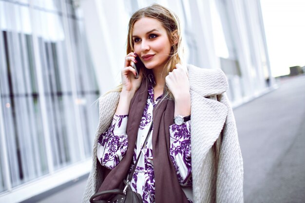 Giovane signora elegante che fa chiamata sul suo smartphone, indossa un cappotto beige alla moda di lusso, sciarpa di cashmere color talpa e abito floreale, in posa vicino al centro business modello.