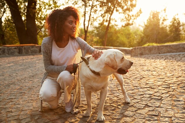 Giovane signora in abbigliamento casual che si siede e che abbraccia cane nel parco