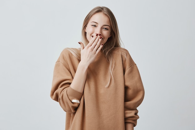 Giovane signora sorridente allegra che copre il viso con la mano. La donna spensierata positiva che indossa i suoi capelli biondi sciolti vide la sorpresa preparata dal marito, in piedi e sorridente, essendo felice